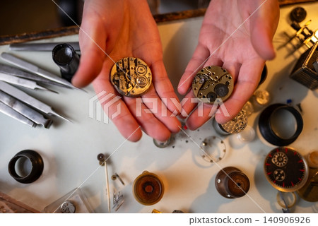 Closeup hands holding vintage mechanical watch movements over repair workspace with tools and parts Closeup hands holding vintage mechanical watch movements over repair workspace with tools and parts 140906926