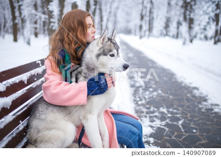 Red-haired girl sitting on a snowy bench with a husky dog Red-haired girl sitting on a snowy bench with a husky dog 140907089