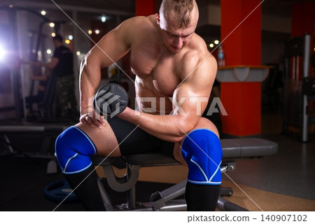 Muscular athlete resting on a gym bench with a heavy dumbbell 140907102