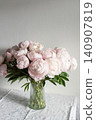 Vertical shot of lush pink peony bouquet with green leaves in ribbed glass vase on table against neutral grey wall in natural light. 140907819