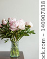 Vertical shot of lush pink peony bouquet with green leaves in ribbed glass vase on dark wooden table against neutral grey wall in natural light. 140907838