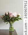 Vertical shot of lush pink peony bouquet with green leaves in ribbed glass vase on dark wooden table against neutral grey wall in natural light. 140907952