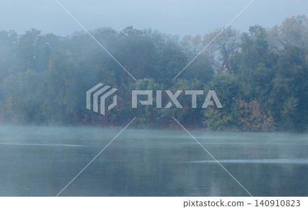 A serene river flows gently through a landscape of trees on a misty autumn morning, creating a tranquil and atmospheric view A serene river flows gently through a landscape of trees on a misty autumn morning, creating a tranquil and atmospheric view 140910823