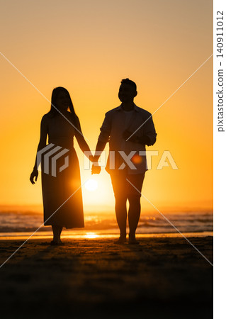 Couple walks hand in hand on the beach during sunset in warm evening light Couple walks hand in hand on the beach during sunset in warm evening light 140911012