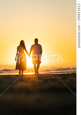 Couple walks hand in hand along the beach at sunset near the ocean in a warm and inviting scene Couple walks hand in hand along the beach at sunset near the ocean in a warm and inviting scene 140911013