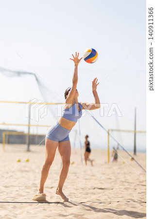 Volleyball player serves ball on beach court while others play in background Volleyball player serves ball on beach court while others play in background 140911366