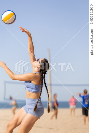 Young woman serves volleyball on sandy beach during bright day with players in background near water Young woman serves volleyball on sandy beach during bright day with players in background near water 140911369