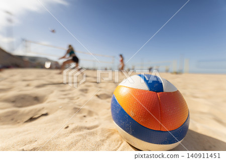 People play beach volleyball while a ball sits in the sand during a sunny day at a coastal location 140911451
