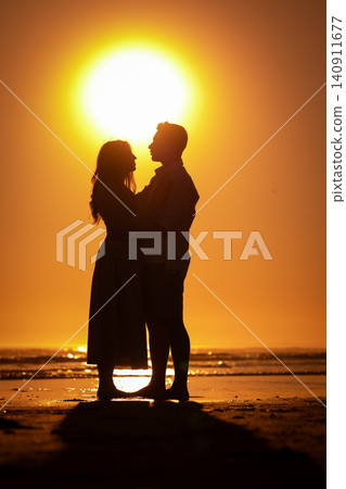 Couple stands together on the beach at sunset with the sun shining behind them Couple stands together on the beach at sunset with the sun shining behind them 140911677
