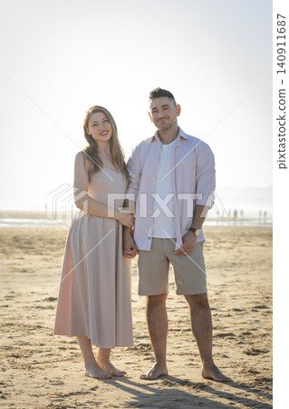 Couple standing hand in hand on a beach during sunset near the shoreline with distant people walking 140911687