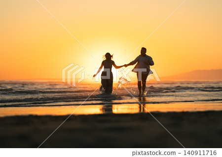 Couple runs along the beach at sunset while splashing in the water near the waves Couple runs along the beach at sunset while splashing in the water near the waves 140911716
