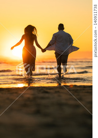 Couple runs hand in hand towards the ocean during sunset on a beach in the evening hours Couple runs hand in hand towards the ocean during sunset on a beach in the evening hours 140911738