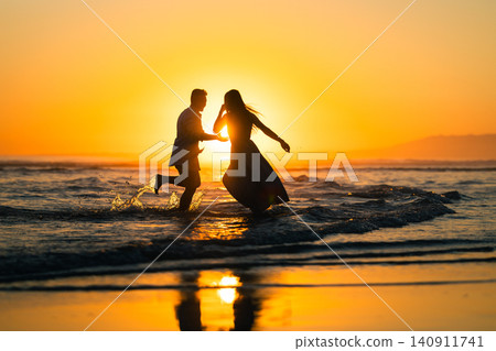 Couple runs along the beach at sunset while waves splash around them and the sun sets on the horizon Couple runs along the beach at sunset while waves splash around them and the sun sets on the horizon 140911741