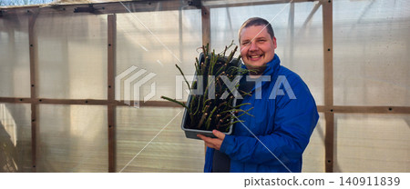 Smiling man holding a tray full of rose stem cuttings ready for propagation and planting in a greenhouse, fostering new growth and developing a horticulture hobby. Smiling man holding a tray full of rose stem cuttings ready for propagation and planting in a greenhouse, fostering new growth and developing a horticulture hobby. 140911839