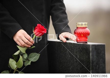 Woman holding red rose near black granite tombstone with candle outdoors, closeup. Funeral ceremony Woman holding red rose near black granite tombstone with candle outdoors, closeup. Funeral ceremony 140911966