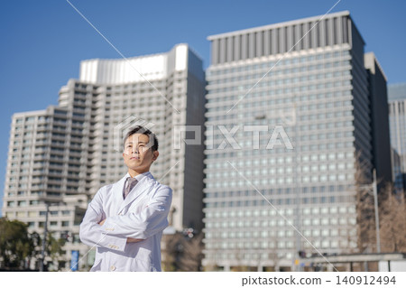A male doctor in a white coat stands with his arms crossed in front of the hospital. A male doctor in a white coat stands with his arms crossed in front of the hospital. 140912494