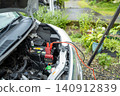 A stock photo of a car helping another car with a dead battery; jumper cables are connected to the battery. 140912839