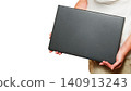 person in a white t-shirt holds a closed black laptop in front of themselves 140913243