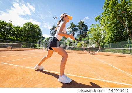Young woman plays tennis on clay court in sunny weather with trees in the background during daytime Young woman plays tennis on clay court in sunny weather with trees in the background during daytime 140913642