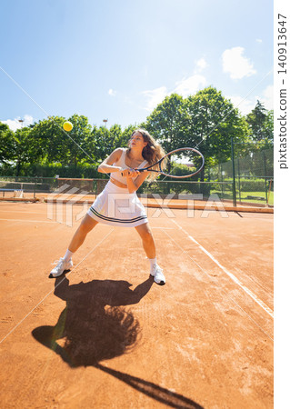 Tennis player hits a ball on a clay court while practicing under a clear sky in the daytime Tennis player hits a ball on a clay court while practicing under a clear sky in the daytime 140913647