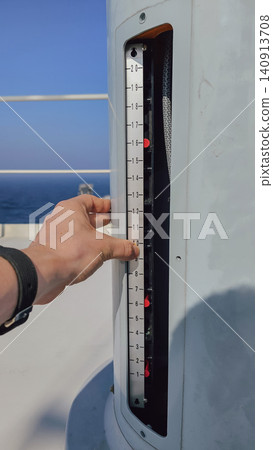 Vertical close up shot of a mariner hand pointing at a metal level indicator scale with numbers inside a column slot on a ship deck, with the blue ocean background Vertical close up shot of a mariner hand pointing at a metal level indicator scale with numbers inside a column slot on a ship deck, with the blue ocean background 140913708