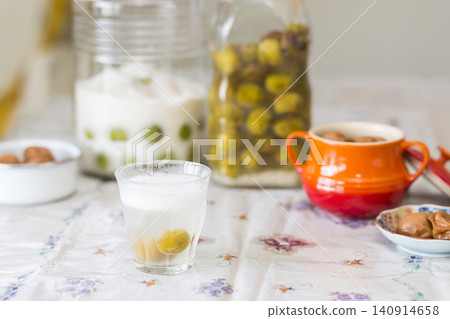 A dining table scene featuring a glass of sparkling water and a background of blurred jars of preserved plums and grapes. 140914658