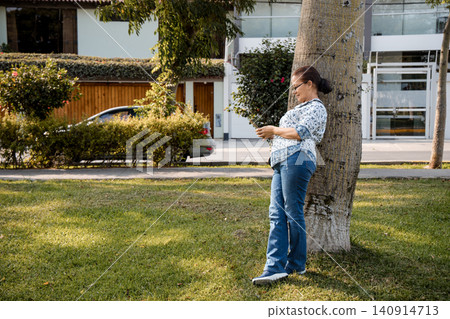 Woman Leaning on Tree and Using Smartphone in Sunny Park Woman Leaning on Tree and Using Smartphone in Sunny Park 140914713