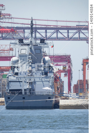 Naval vessel docked at busy port with cargo cranes in background during bright daylight 140914884