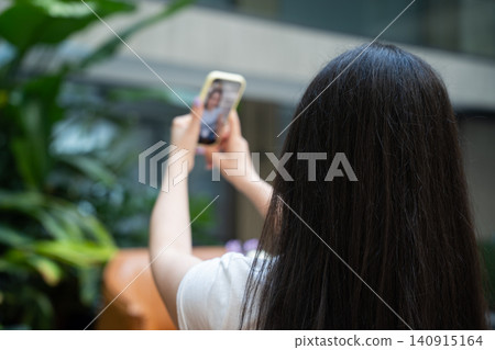 Rear view of young woman taking a smartphone photo in a modern indoor space with lush green plants. Travel photography mobile photography and content creator lifestyle with smartphone Rear view of young woman taking a smartphone photo in a modern indoor space with lush green plants. Travel photography mobile photography and content creator lifestyle with smartphone 140915164