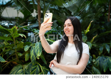 Young woman taking a selfie with a smartphone in a modern indoor space surrounded by lush green plants. Social media content creator lifestyle and personal branding Young woman taking a selfie with a smartphone in a modern indoor space surrounded by lush green plants. Social media content creator lifestyle and personal branding 140915170