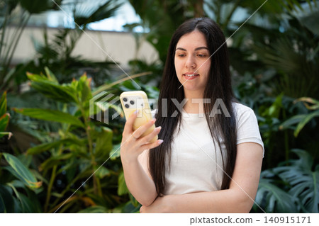 Young woman using a smartphone in a modern indoor space with lush green plants. Mobile technology digital communication and mobile app use for everyday lifestyle Young woman using a smartphone in a modern indoor space with lush green plants. Mobile technology digital communication and mobile app use for everyday lifestyle 140915171