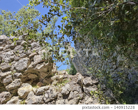 Ruins of a medieval fortress wall stand tall amid green trees in the Spanish countryside. Sunlight highlights the stone structure, revealing its ancient history. Ruins of a medieval fortress wall stand tall amid green trees in the Spanish countryside. Sunlight highlights the stone structure, revealing its ancient history. 140915228