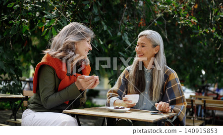 Two Mature Women Friends Sharing Warm Conversation Over Coffee at a Sunlit Outdoor Cafe Table Surrounded by Lush Green Trees Two Mature Women Friends Sharing Warm Conversation Over Coffee at a Sunlit Outdoor Cafe Table Surrounded by Lush Green Trees 140915853