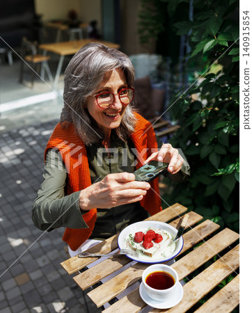 Stylish Mature Woman Photographing Her Strawberry Dessert with a Smartphone at a Sunny Outdoor Cafe Table Beside Green Foliage Stylish Mature Woman Photographing Her Strawberry Dessert with a Smartphone at a Sunny Outdoor Cafe Table Beside Green Foliage 140915854