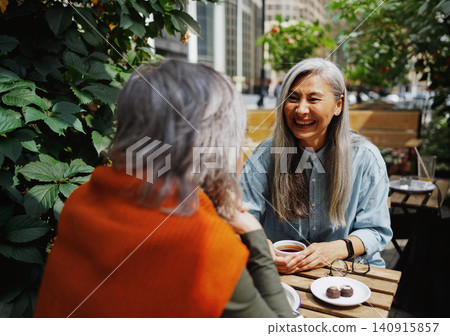 Authentic Mature Asian Woman Laughing Openly with a Friend Over Tea and Chocolates at a Green Urban Outdoor Cafe in Soft Daylight Authentic Mature Asian Woman Laughing Openly with a Friend Over Tea and Chocolates at a Green Urban Outdoor Cafe in Soft Daylight 140915857