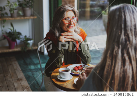 Engaged Mature Woman in Red Glasses Listening Warmly to a Friend at a Cozy Indoor Cafe Table with Tea and Strawberry Toast Engaged Mature Woman in Red Glasses Listening Warmly to a Friend at a Cozy Indoor Cafe Table with Tea and Strawberry Toast 140915861