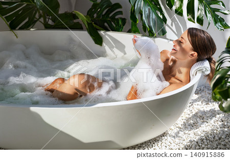 Happy young Caucasian woman relaxing in a bubble bath inside a bright modern bathroom Happy young Caucasian woman relaxing in a bubble bath inside a bright modern bathroom 140915886