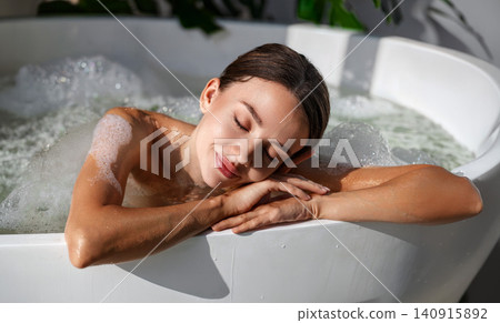 Relaxed young Caucasian woman resting head on arms while taking a warm bubble bath in bathtub 140915892