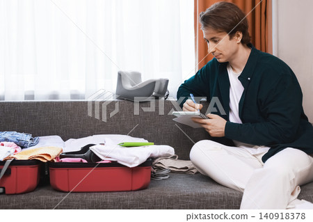 Man preparing luggage for a vacation trip, writing a packing list in a notebook. He sits on a sofa next to an open red suitcase filled with clothes. Man preparing luggage for a vacation trip, writing a packing list in a notebook. He sits on a sofa next to an open red suitcase filled with clothes. 140918378