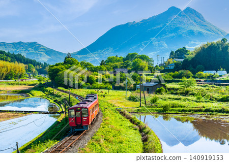 The KiHa 200 series train runs through the early summer countryside with Mount Yufu in the background on the Kyudai Main Line. 140919153