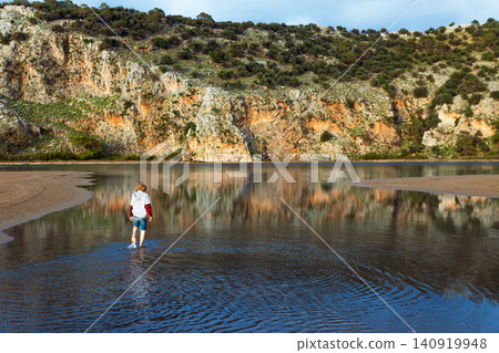 Young boy walks through the shallow lagoon at Iztuzu Beach near Dalyan, Turkey. Peaceful family travel moment surrounded by water, cliffs and protected coastal nature during summer vacation. Young boy walks through the shallow lagoon at Iztuzu Beach near Dalyan, Turkey. Peaceful family travel moment surrounded by water, cliffs and protected coastal nature during summer vacation. 140919948