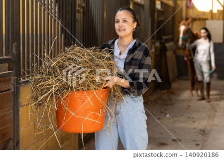 Asian young female worker walks go through corridor of stable, carrying basket with hay 140920186
