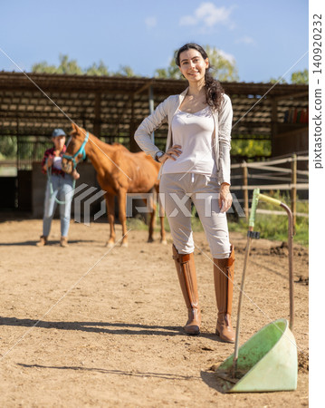 Armenian female stable worker stands near wooden fence of paddock and watches horse. 140920232