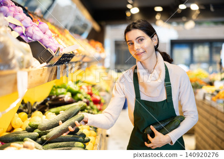 Young woman seller puts cucumbers in vegetable shop 140920276