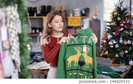 Woman stands in front of a Christmas tree and chooses a green reindeer sweater Woman stands in front of a Christmas tree and chooses a green reindeer sweater 140920332