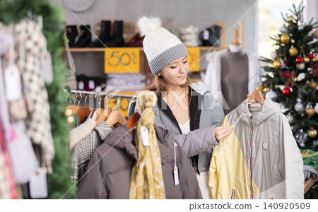 Girl client looks at products in store, looks closely at down jacket, picks up wardrobe item Girl client looks at products in store, looks closely at down jacket, picks up wardrobe item 140920509