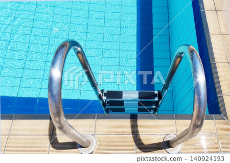 An overhead view of a staircase with stainless steel railings leading into the clear blue water of a pool with tiled floors and walls. The concept is for swimming, summer relaxation, and recreation. An overhead view of a staircase with stainless steel railings leading into the clear blue water of a pool with tiled floors and walls. The concept is for swimming, summer relaxation, and recreation. 140924193