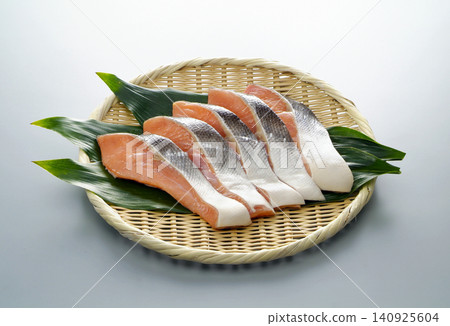 A photograph of slices of autumn salmon arranged on a bamboo basket lined with haran leaves, against a white to gray background. A photograph of slices of autumn salmon arranged on a bamboo basket lined with haran leaves, against a white to gray background. 140925604