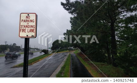 A no-parking, fire lane sign along a wet rural road on a rainy day. A paved sidewalk runs alongside the roadway, near a forested area, with no people. A no-parking, fire lane sign along a wet rural road on a rainy day. A paved sidewalk runs alongside the roadway, near a forested area, with no people. 140925748