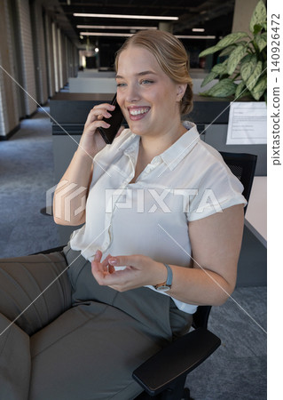 Woman smiling, holding phone to right ear while gesturing from chair in office wearing white blouse Woman smiling, holding phone to right ear while gesturing from chair in office wearing white blouse 140926472